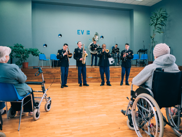Eine Gruppe von Menschen im Rollstuhl, die in einem Raum mit einer Wand im Hintergrund, Pflanzen auf beiden Seiten und Lampen an der Decke Musikinstrumente spielen.