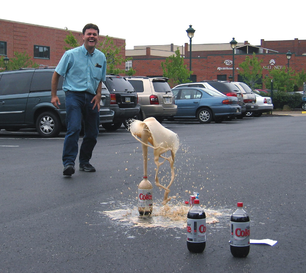 Ein Mann steht auf einer Straße, lacht und hat drei Flaschen vor sich, während im Hintergrund Autos, eine Pflanze, ein Lichtmast und der Himmel zu sehen sind.