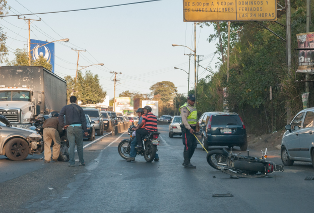 Gruppe von Menschen, die um ein verunglücktes Motorrad auf der Seite einer Straße mit mehreren Fahrzeugen, Bäumen, Strommästen und einem klaren Himmel im Hintergrund stehen.