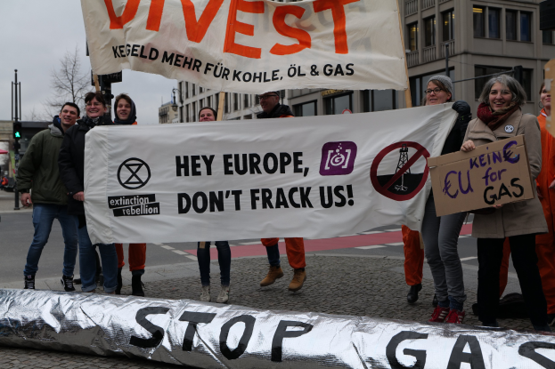 Gruppe von Menschen mit einem "Stoppt Gas"-Schild auf einer Straße mit Gebäuden, Bäumen, Verkehrsschildern im Hintergrund unter einem klaren blauen Himmel.