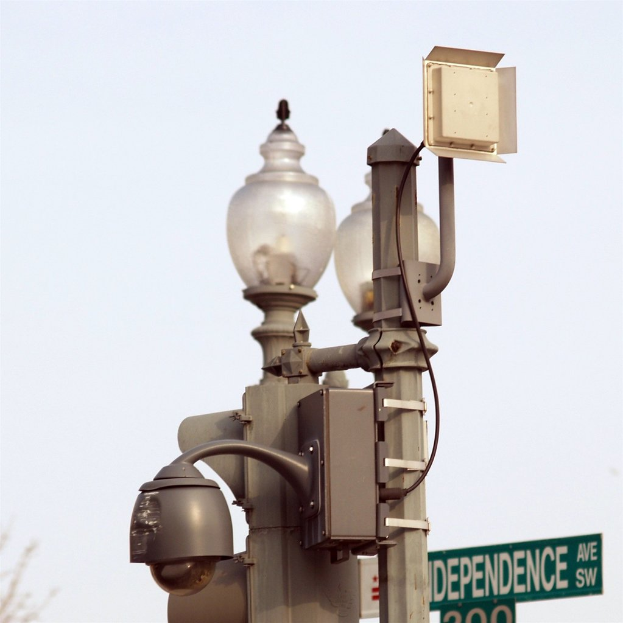 Zwei Pfosten, einer mit Kamera und Licht, der andere mit Lichtern, vor einem weißen Himmel, mit einem Schild unten.