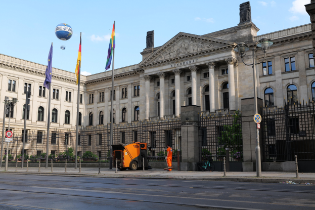 Großes modernes Gebäude mit vielen Fenstern als Deutscher Bundestag in Berlin identifiziert, umgeben von Straßeninfrastruktur, Fußgängern, Flaggen und Bäumen unter einem bewölkten Himmel.