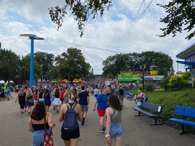 Eine Menschenmenge, die eine Straße mit Bänken, Pflanzen, Gras, Bäumen, Pfosten, Lampen, Drähten und einem Schuppen gesäumt, mit einigen, die Taschen tragen, unter einem bewölkten Himmel.