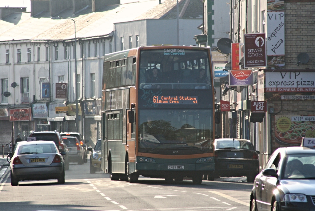 Eine Straße mit Autos und einem Bus im Vordergrund, mit Gebäuden mit Wänden, Fenstern, Tellern und Dächern im Hintergrund, sowie Plakaten und Bannern an den Wänden und einem Laternenmast.