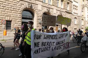 Eine Gruppe von Menschen, die während einer Demonstration in Berlin durch die Straßen gehen, einige halten Schilder und Banner, andere fahren Fahrräder, im Hintergrund ein Gebäude mit Fenstern, Bögen, Säulen und Skulpturen.