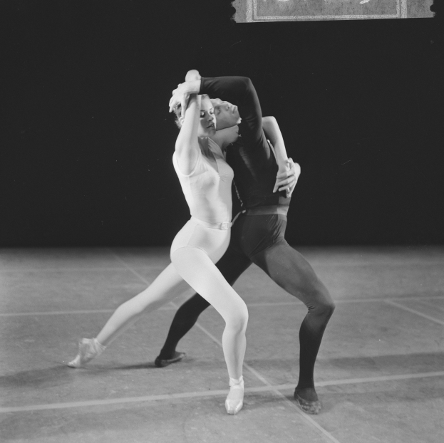 Ein Schwarz-Weiß-Foto eines Mannes in einem weißen Hemd und schwarzen Hosen und einer Frau in einem unbekannten Outfit, die anmutig und perfekt synchron auf einer Bühne tanzen.