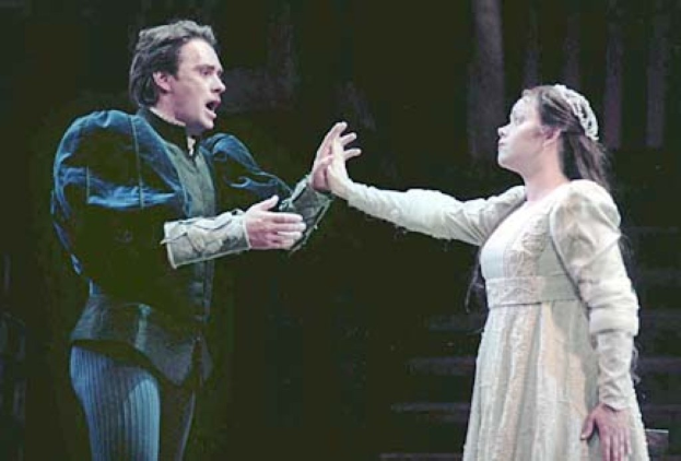 Ein Mann in einem schwarzen Anzug und weißem Hemd und eine Frau in einem langen, wallenden Kleid stehen auf einer Bühne und machen ernste Gesichter, wobei der Mann ein Objekt hält und eine dunkle Treppe im Hintergrund zu sehen ist.