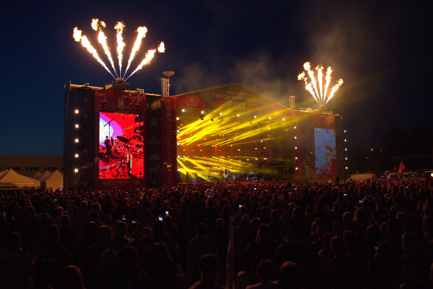 Eine große Menschenmenge auf einem Musikfestival schaut auf einer hell erleuchteten Bühne mit Bannern, umgeben von Zelten und Feuerwerk am Nachthimmel.