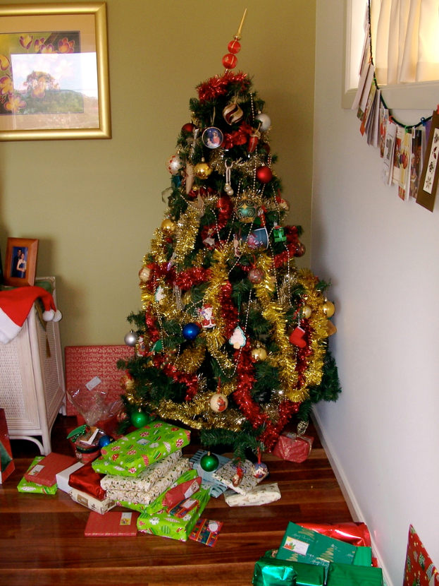 Ein geschmückter Weihnachtsbaum rechts mit Geschenken darunter, ein Foto Rahmen an der linken Wand und ein Vorhang am rechten Fenster.