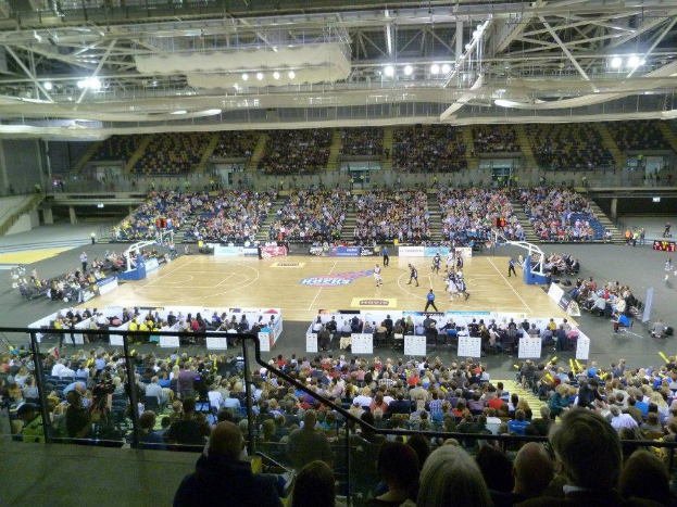 Eine große Menschenmenge in einer Arena, die ein Basketballspiel schaut, mit Texttafeln rund um das Feld und einer Decke mit Stangen und Lampen darüber.