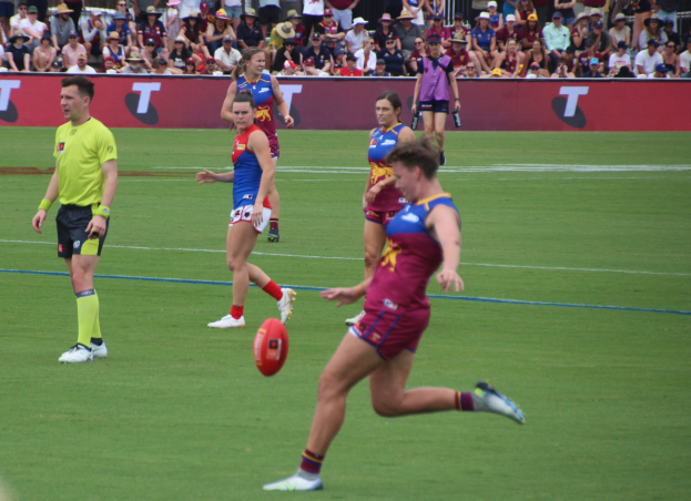 Eine Gruppe von Menschen, die Australian Football auf einem grünen Feld spielen, mit einem Ball in der Mitte der Aktion und Zuschauern und Geländern im Hintergrund.