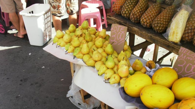 Eine Marktszene mit verschiedenen Fruächten auf einem Gestell, einigen Hockern, einigen Menschen und zwei Preisplakaten.