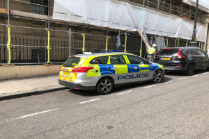 Ein Polizeiwagen steht auf der Straße neben einem Gebäude, zwei Helme tragende Personen stehen daneben; im Vordergrund sind ein Geländer und ein Pfahl zu sehen, im Hintergrund ein weißes Tuch über das Gebäude drapiert.