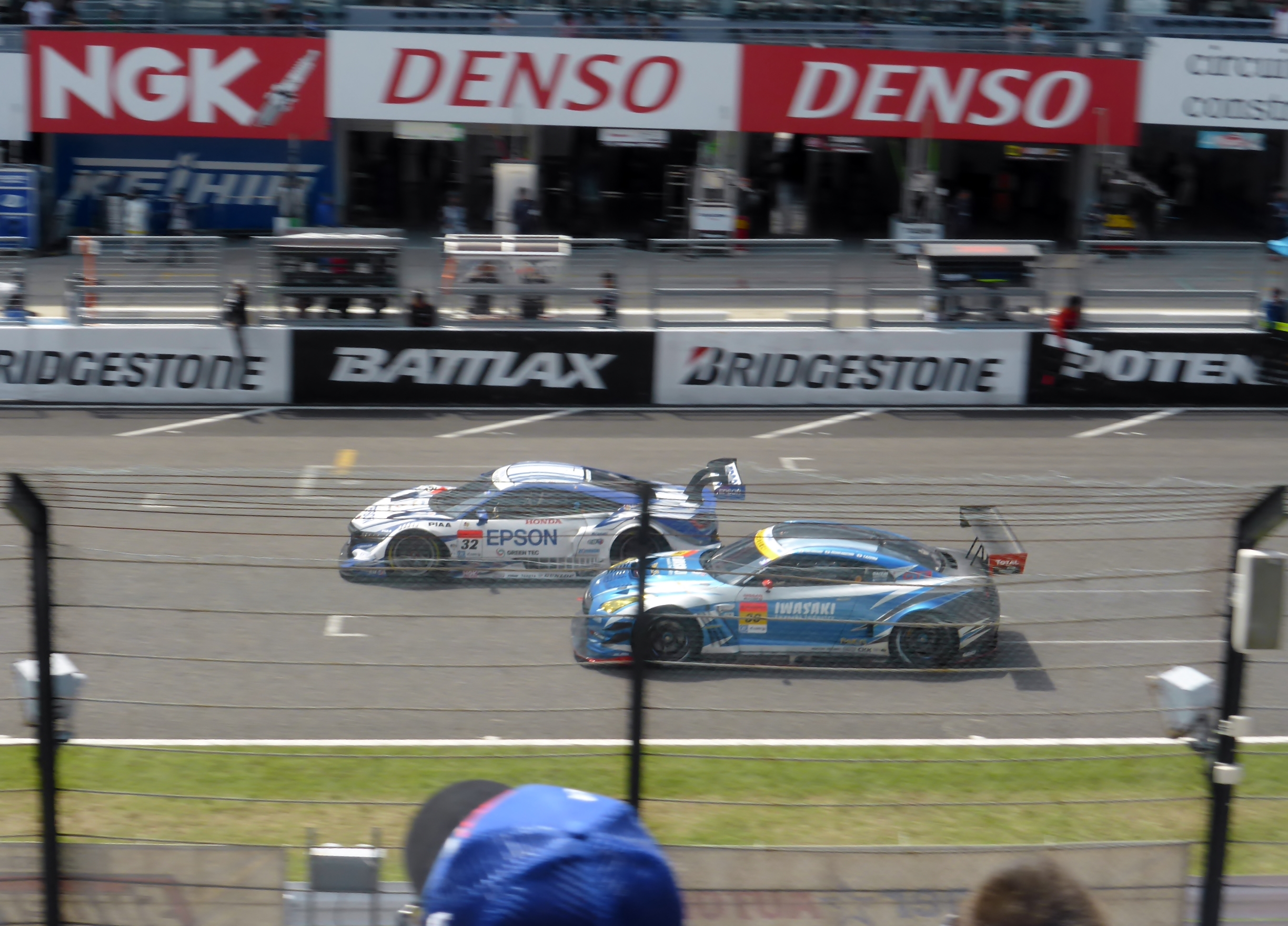 Zwei Sportwagen rennen auf einer Rennstrecke vor einer Zuschauermenge hinter einem Maschendrahtzaun, Pfosten und Lichtern, mit Texttafeln im Hintergrund.