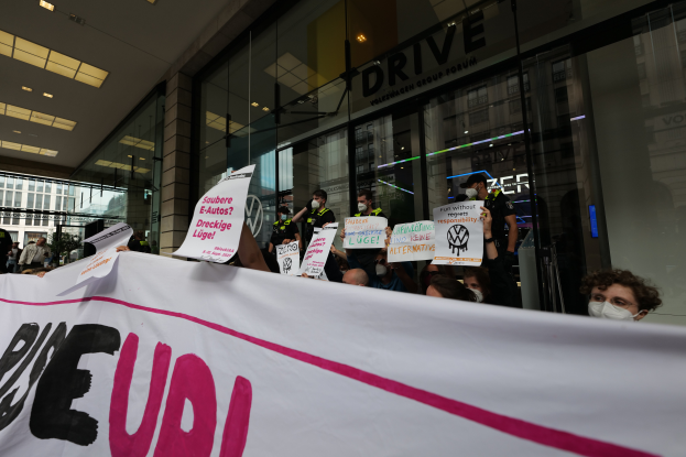 Eine Gruppe maskierter Personen mit einer Fahne, auf der 'Volkswagen Gruppe' steht, protestiert vor einem Glasgebäude.