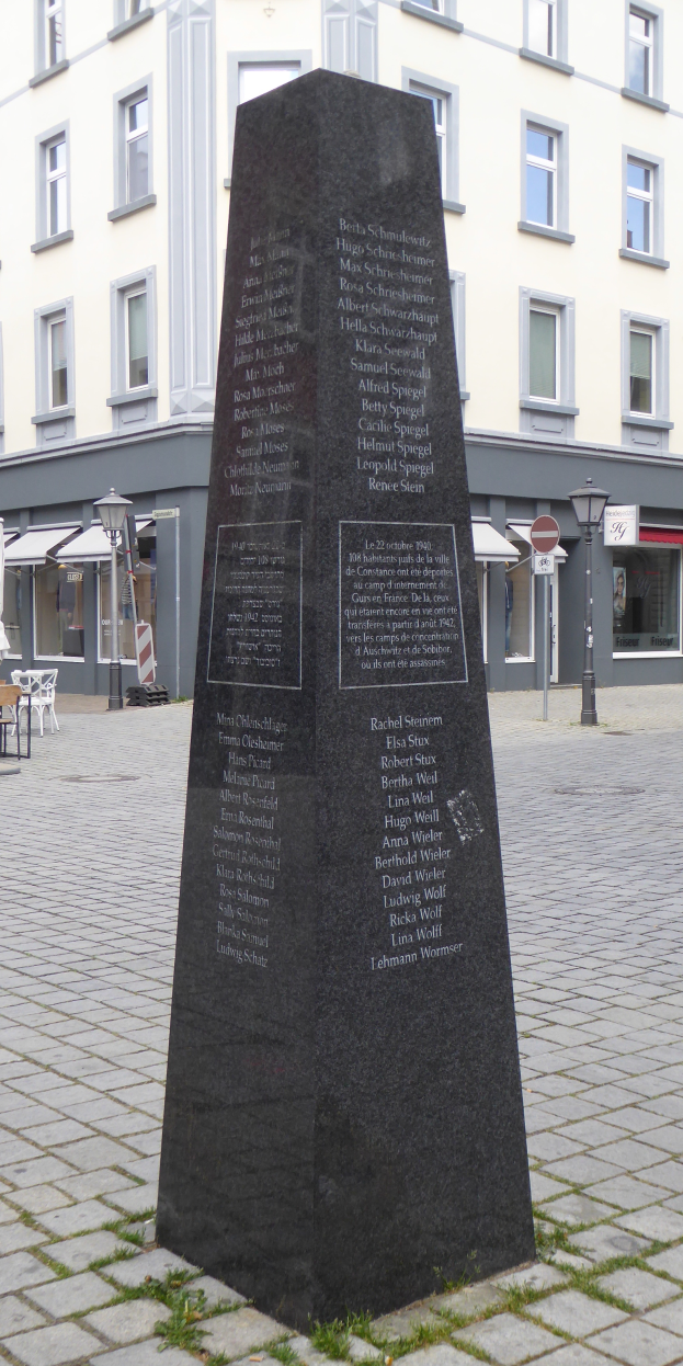Ein Denkmal mit Inschrift steht auf einem Stadtplatz, umgeben von Gebäuden, Straßenlaternen, Schildern, Tischen, Stühlen und Menschen, mit dem Himmel im Hintergrund.