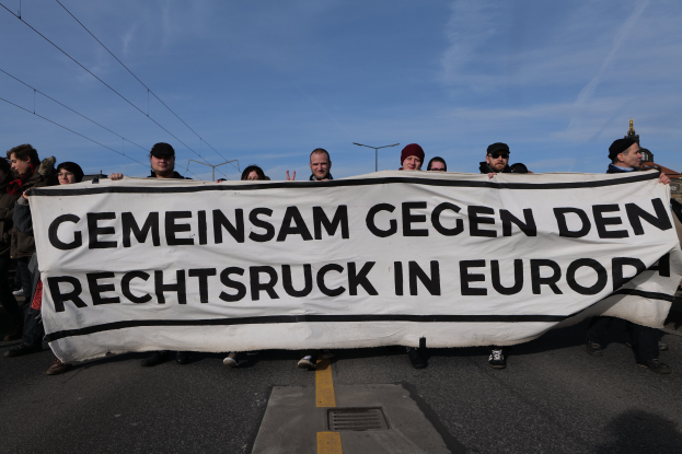 Eine Gruppe von Menschen steht auf der Straße und hält ein Transparent mit der Aufschrift "Gemeinsam Gegen den Rechtsruck in Europa" in den Händen mit Straßenlaternen, Strommasten, Stromkabeln, Gebäuden und einem bewölkten Himmel im Hintergrund.
