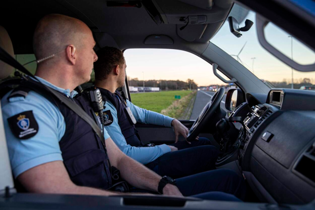 Zwei Polizeibeamte sitzen im Fahrerhaus eines Autos auf einer von Gras, Bäumen und Masten umgebenen Straße, mit einem klaren blauen Himmel im Hintergrund.