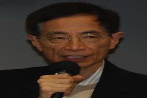 Ein Mann in einem schwarzen Blazer und Brille hält ein Mikrofon und sieht ernst aus, wahrscheinlich ein Ökonom.