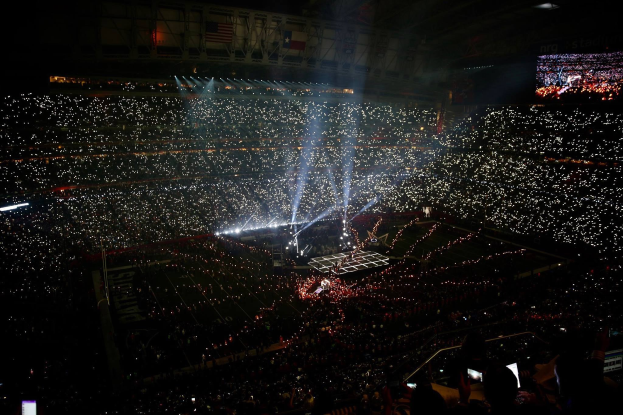 Eine große Menschenmenge in einem hell erleuchteten Stadion bei Nacht mit einem Bildschirm auf der rechten Seite, der die Ergebnisse der Super Bowl XLVII-Halbzeitshow anzeigt.