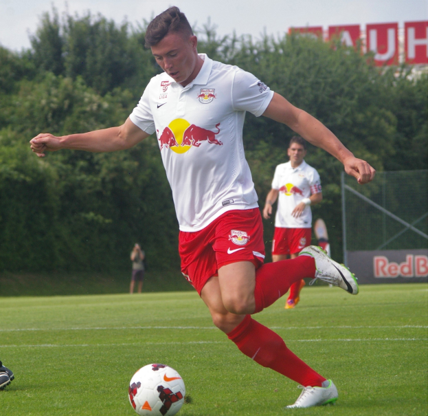Ein professioneller Fußballspieler in einem roten Trikot schießt einen Fußball auf einem grünen Feld mit Bäumen und einem klaren blauen Himmel im Hintergrund, wobei "RB Leipzig" auf einem Schild zu sehen ist.