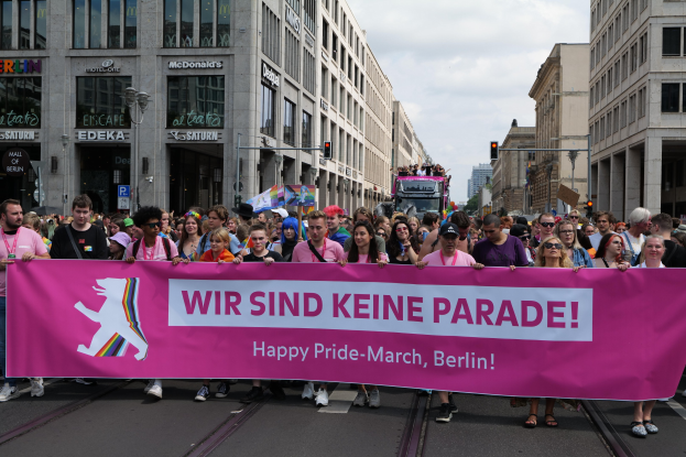 Eine Gruppe von Menschen, die eine rosa Schriftzug-Tafel mit der Aufschrift "Happy Pride March" trägt, geht eine Straße in Berlin, Deutschland, entlang, mit Gebäuden, Laternenmasten und Verkehrszeichen an der Straße und einer bewölkten Himmel im Hintergrund.