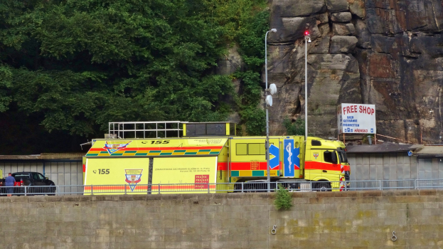 Ambulanz am Rande einer Straße neben einer Klippe geparkt, mit verschiedenen Straßenobjekten im Vordergrund und Bäumen und einem Auto im Hintergrund.