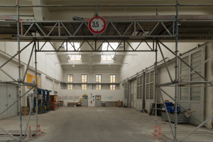 Ein großes Lager mit ausgedehnten Gerüsten, Verkehrskegeln auf dem Boden, Metallstangen, einem Schild, verschiedenen Maschinen, einer Tür, Fenstern, einer Wand und einem Dach mit Deckenbeleuchtung.