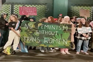 Eine Gruppe von Menschen hält ein Transparent mit der Aufschrift "Kein Feminismus ohne trans Frauen" und Schilder in einer öffentlichen Fläche mit einer Wand und einem Rollladen im Hintergrund.