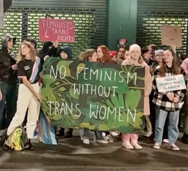Eine Gruppe von Menschen hält ein Transparent mit der Aufschrift "Kein Feminismus ohne trans Frauen" und Schilder in einer öffentlichen Fläche mit einer Wand und einem Rollladen im Hintergrund.