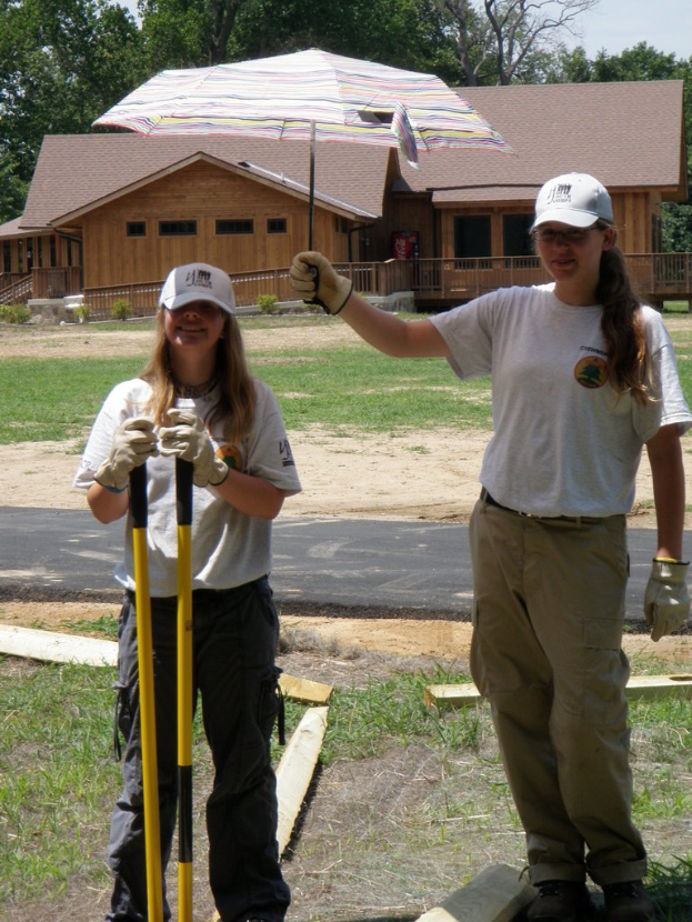 Zwei Frauen in Mützen und Handschuhen stehen auf einer grasigen Fläche neben einer Straße; eine hält einen Schirm, die andere Stöcke, mit einem Gebäude und Bäumen im Hintergrund.