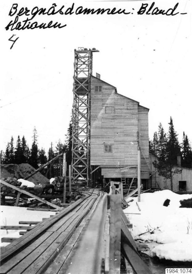 Ein Schwarz-Weiß-Foto einer Bahntrasse mit einem Gebäude im Hintergrund, umgeben von Bäumen, Pfählen und Schnee, mit dem Text "Berghäsdammen Bland 4" oben und unten.
