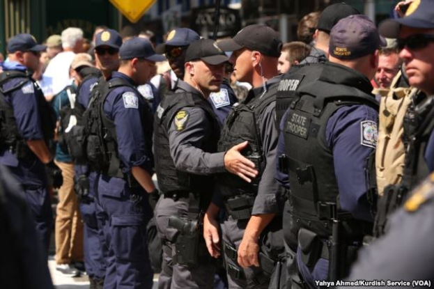 Eine Gruppe von Polizisten in Uniform steht in einer Reihe auf einer Straße, einige halten Feuerwaffen, mit Gebäuden und Beschriftungen im Hintergrund.
