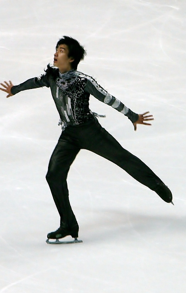 Männlicher Eiskunstläufer in schwarzer Kleidung gleitet über eine Eisbahn und führt eine Choreografie mit synchronen Arm- und Beinbewegungen aus.