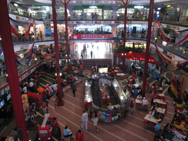 Ein belebtes Einkaufszentrum-Innere mit Säulen, Menschen, Tischen, Fliesen, Flaggen, Geschäften, einem Banner, Treppen und einem Namensschild.
