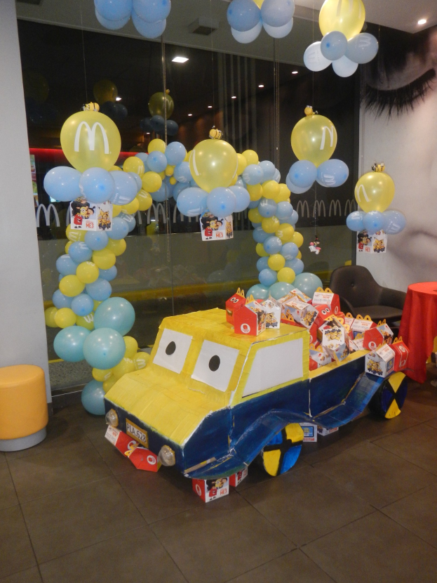 Eine McDonald's-Geburtstagsfeier mit einem gelben und blauen Lastwagen, der mit Ballons und anderen Dekoartikeln geschmückt ist, einem rot gedeckten Tisch mit Gegenständen, Hockern und einer Glaswand im Hintergrund unter festlicher Deckenbeleuchtung.