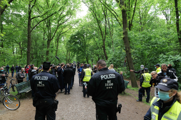 Eine Gruppe von Polizisten steht vor einer Menschenmenge bei einem Outdoor-Event, mit Fahrrädern und einer Bank im Vordergrund und Bäumen im Hintergrund.