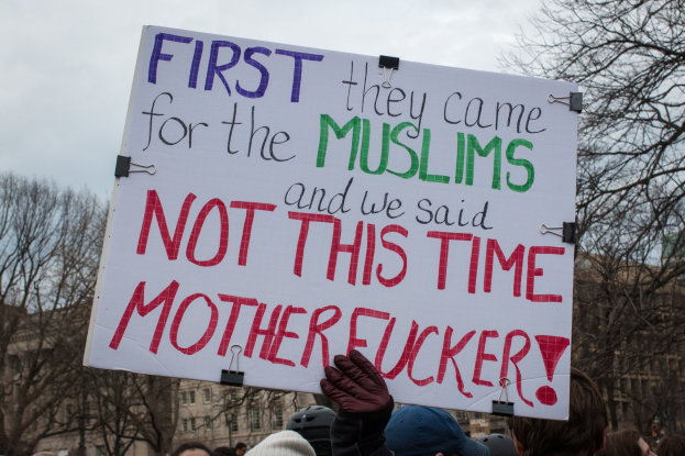Eine Gruppe von Menschen mit Möppen und Handschuhen hölt ein Schild mit der Aufschrift "Zuerst kamen sie wegen der Muslime und wir sagten nicht dieses Mal Arschloch" mit Bäumen, Gebäuden und einem klaren blauen Himmel im Hintergrund.