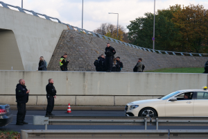 Eine Gruppe von Polizisten steht neben einem Auto auf der Straße, mit Verkehrskegeln, einer Barriere, Gras, einer Wand, Laternen, Bäumen und einem bewölkten Himmel im Hintergrund.