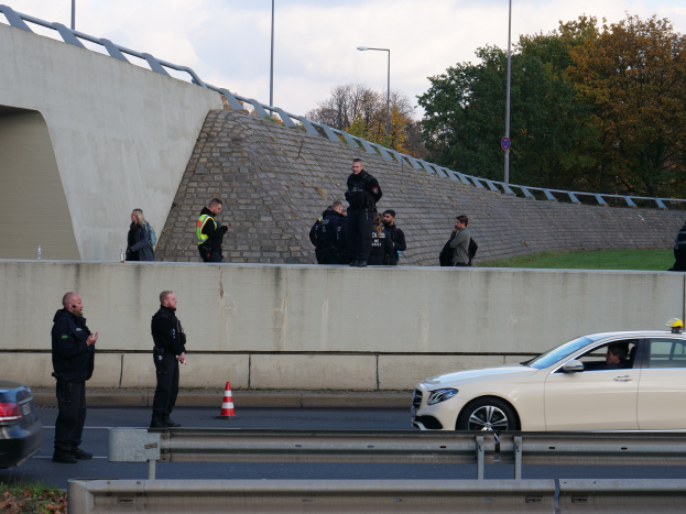 Eine Gruppe von Polizisten steht neben einem Auto auf der Straße, mit Verkehrskegeln, einer Barriere, Gras, einer Wand, Laternen, Bäumen und einem bewölkten Himmel im Hintergrund.
