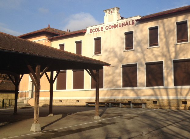 Ein Schulgebäude mit einem Schild, das "Ecole Communale" trägt, vorne, mit einem Schuppen mit Säulen, Bänken, Treppen mit Geländern, Bäumen und einem bewölkten Himmel.