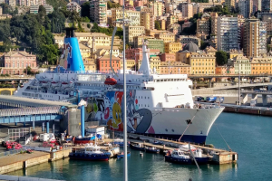 Ein großes Kreuzfahrtschiff liegt nahe einer Stadt mit einer Brücke auf der linken Seite, Fahrzeugen auf der Straße und Gebäuden und Bäumen im Hintergrund.