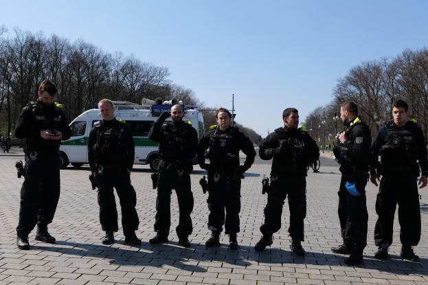 Eine Gruppe von Polizisten steht vor einem Rettungswagen auf einer Straße mit Bäumen, Laternenmästen und einem klaren Himmel.