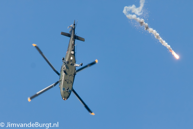 Ein militärisches Hubschrauber in der Luft, das eine Rakete hält, die Rauch und Feuer ausstößt, mit Text in der unteren linken Ecke.