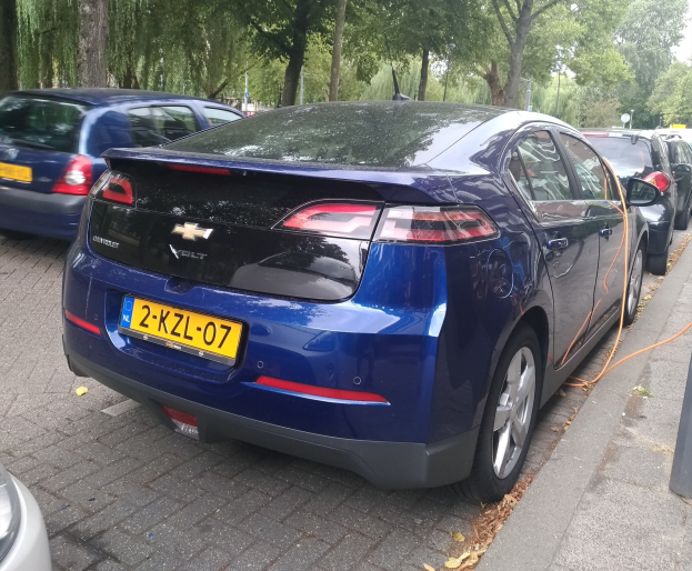 Elektrisches Auto an einer Ladestation auf einer Straße mit Bäumen unter einem klaren blauen Himmel.
