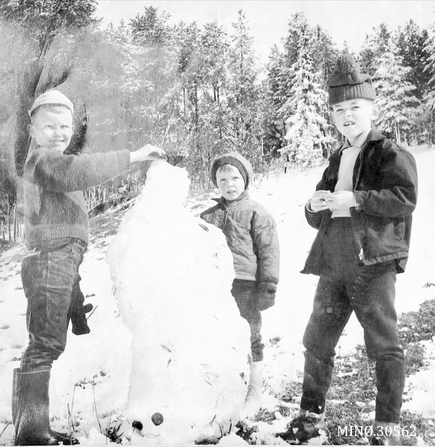 Drei junge Jungs in Jacken und Mützen bauen einen Schneemann in einer verschneiten Landschaft mit schneebedeckten Bäumen, dargestellt in Schwarz-Weiß.