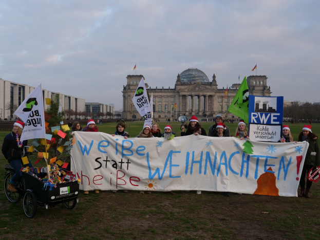 Eine Gruppe von Menschen mit Mützen, die ein Banner vor dem Reichstaggebäude in Berlin, Deutschland, halten, mit einer Person auf einem Kinderwagen, grasbewachsenem Boden, Bäumen, Gebäuden, Flaggen und einem bewölkten Himmel im Hintergrund.
