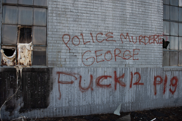 Ein Gebäude mit einer Graffiti-Seite, auf der "Polizei Mord George Fuck 12 P19" steht, mit Fenstern und verschiedenen Gegenständen davor.