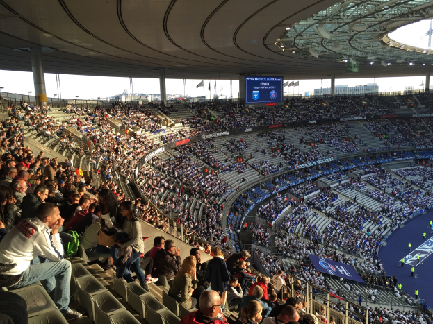 Große Menschenmenge in einem Stadion bei einem Füßtballspiel mit einer Bühne, Fahnen, Masten, einem Bildschirm und der Allianz Arena in München, Deutschland im Hintergrund.
