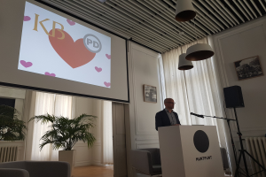 Mann an einem Rednerpult stehend und in ein Mikrofon sprechend, mit einem projizierten Bildschirm dahinter, umgeben von Stühlen, einer Pflanze, Fotorahmen, Vorh├Ąngen und Deckenleuchten.